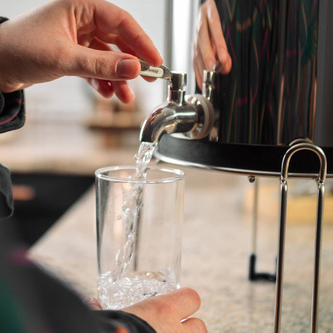 Hand opening a polished stainless steel water spigot to fill a clear glass with filtered water from a raised gravity water filter stand, featured in Appliances and Emergency Supplies collections.
