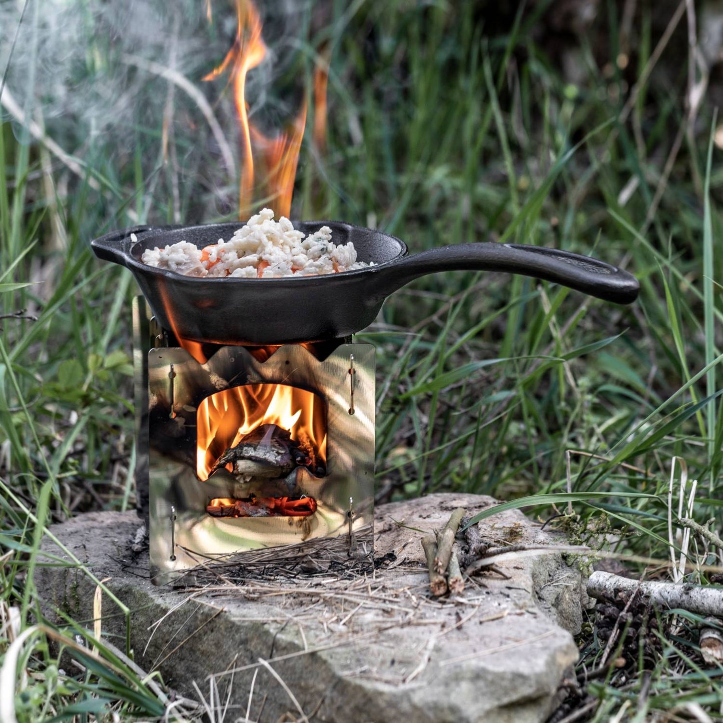 Cast iron skillet with long handle cooking food over an open flame on a compact portable stove, ideal for camping, kitchen, and outdoor use, featured in Farm, Garden & Tool and Cookware collections.