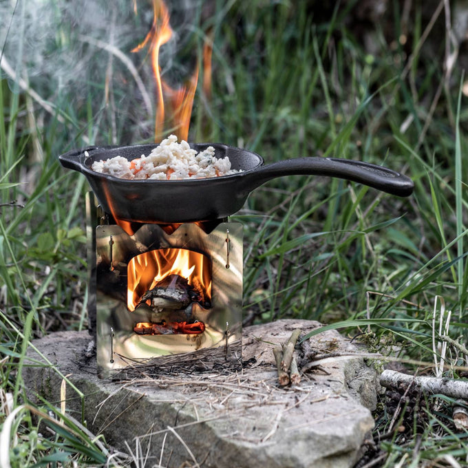 Cast iron skillet with long handle cooking food over an open flame on a compact portable stove, ideal for camping, kitchen, and outdoor use, featured in Farm, Garden & Tool and Cookware collections.