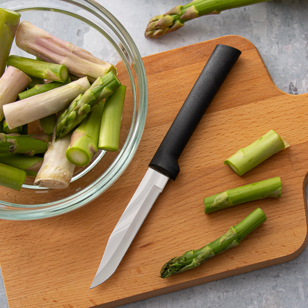Handcrafted serrated paring knife with a finely toothed stainless steel blade and smooth black handle, made in USA, shown slicing fresh asparagus on a wooden cutting board, ideal for Farm, Garden & Tool and Kitchen & Food collections.
