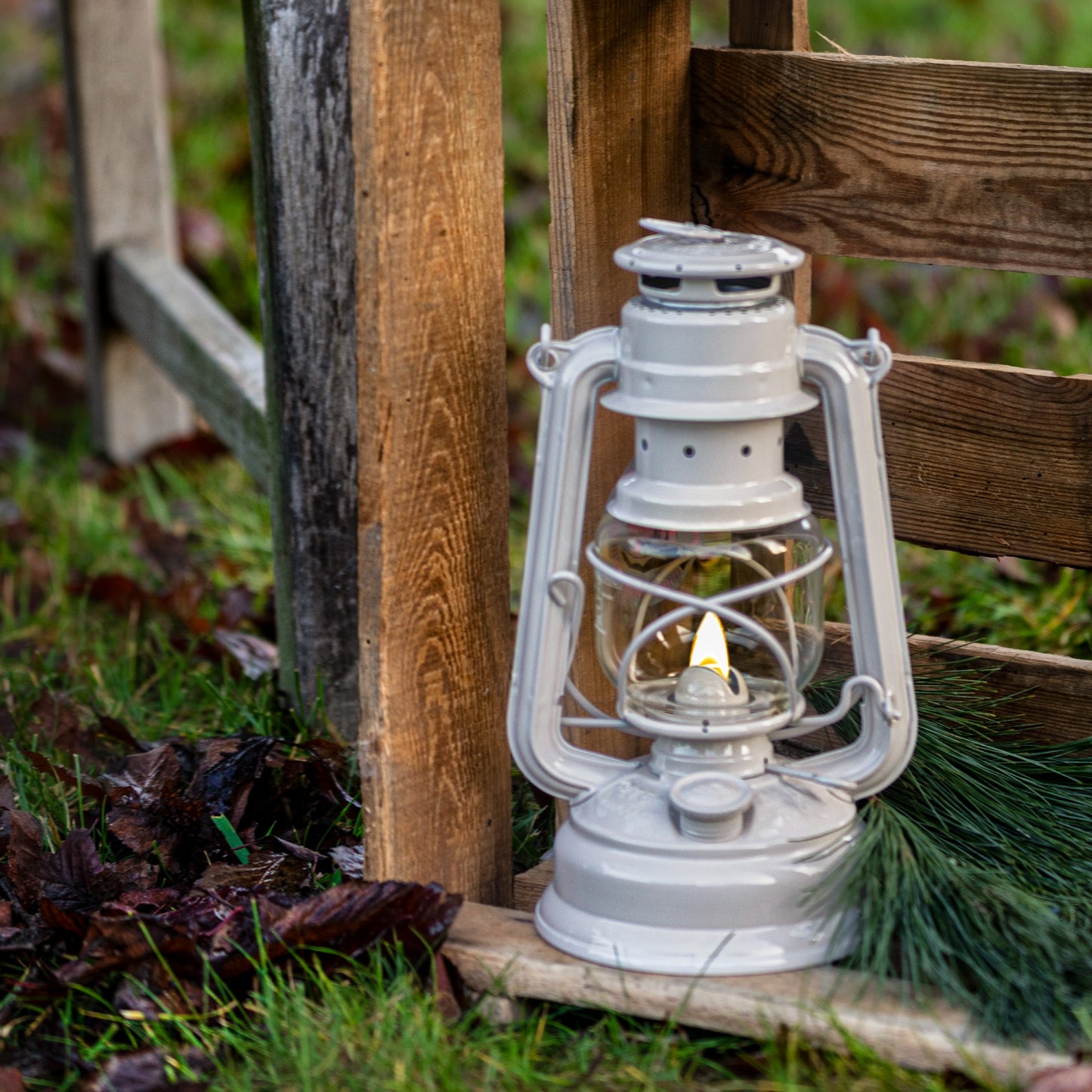 Handcrafted galvanized oil lantern with thermal heat-resistant globe softly glowing beside a rustic wooden fence on damp grass with pine sprigs, ideal for Camping and Emergency Supplies.