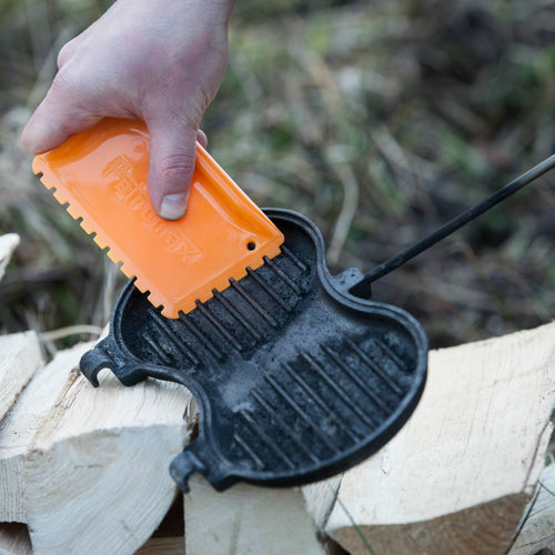 Handheld orange scraper cleaning a cast iron burger iron with ridged round molds, used for outdoor cooking in camping, cookware, and farm tool collections.