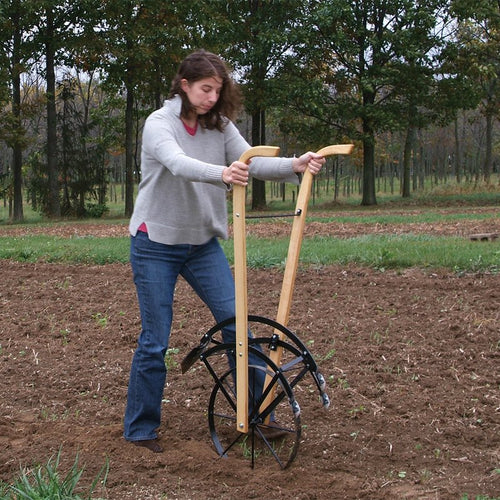 Woman using a handcrafted Amish-made cultivator with wooden handles and a solid steel frame to till soil in a garden, shown in an outdoor farm setting during fall.