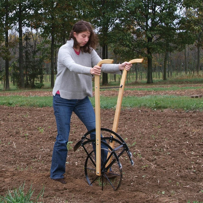Woman using a handcrafted Amish-made cultivator with wooden handles and a solid steel frame to till soil in a garden, shown in an outdoor farm setting during fall.