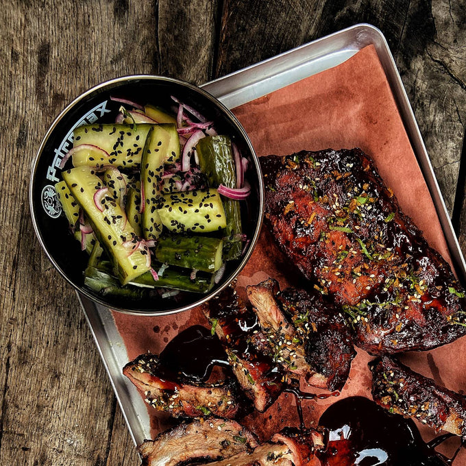 Black enamel bowl filled with pickled cucumbers and red onions, placed on a metal tray with grilled ribs garnished with herbs, featured in Kitchen & Food, Camping, Dinnerware, and Best Sellers collections.