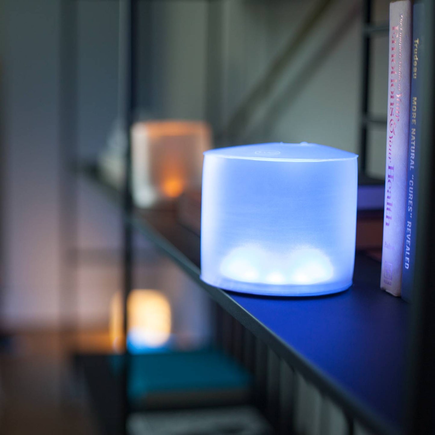 Soft blue glowing Luci Color Essence solar light on a shelf next to books, adding colorful, eco-friendly illumination for camping, emergency supplies, and garden lighting.