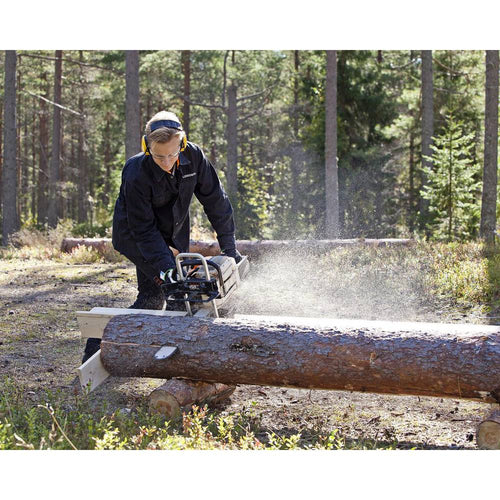 Man wearing protective headphones and dark clothing uses a portable sawmill attachment on a chainsaw to slice a large log outdoors in a forest clearing, featured in Farm, Garden & Tool and Wood Cutting and Hauling collections.