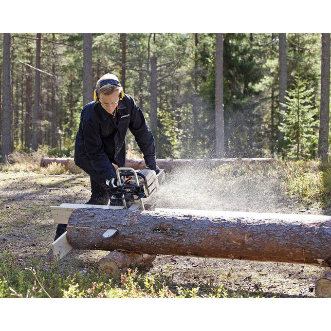 Man wearing protective headphones and dark clothing uses a portable sawmill attachment on a chainsaw to slice a large log outdoors in a forest clearing, featured in Farm, Garden & Tool and Wood Cutting and Hauling collections.