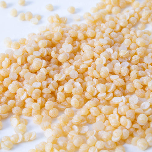 Close-up of light yellow natural beeswax pellets scattered on a white surface, handcrafted for use in candle making and featured in lamps, candlemaking supplies, and best sellers collections.