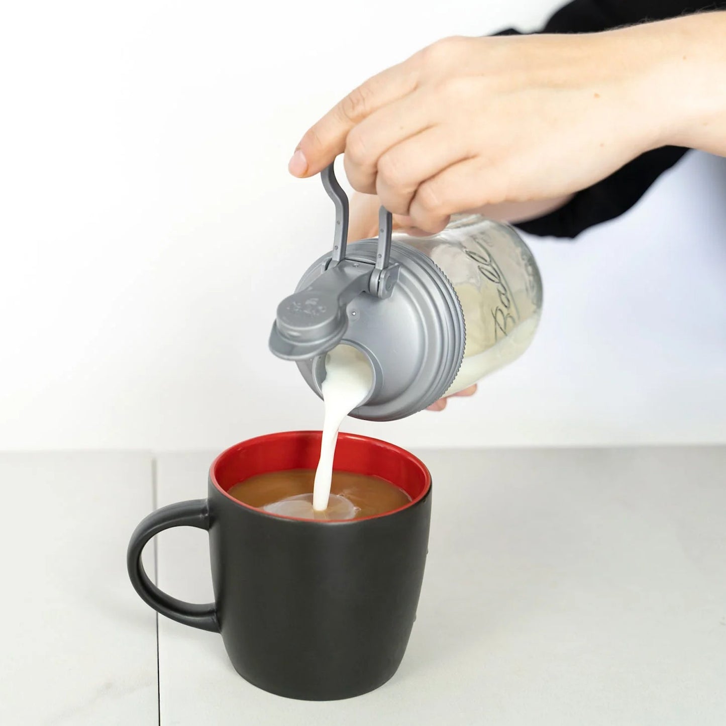 Hand pouring milk from a clear glass jar with a gray no-break plastic pour lid featuring a flip-top spout and carry loop into a black coffee mug, showcasing durable USA-made kitchen accessories ideal for Kitchen & Food, Dinnerware, Kitchen Gadgets, Kitchen Accessories, Best Sellers, and Promo Eligible collections.