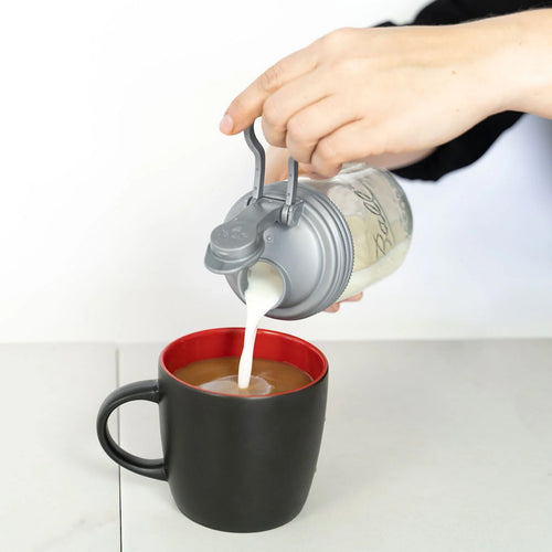 Hand pouring milk from a clear glass jar with a gray no-break plastic pour lid featuring a flip-top spout and carry loop into a black coffee mug, showcasing durable USA-made kitchen accessories ideal for Kitchen & Food, Dinnerware, Kitchen Gadgets, Kitchen Accessories, Best Sellers, and Promo Eligible collections.