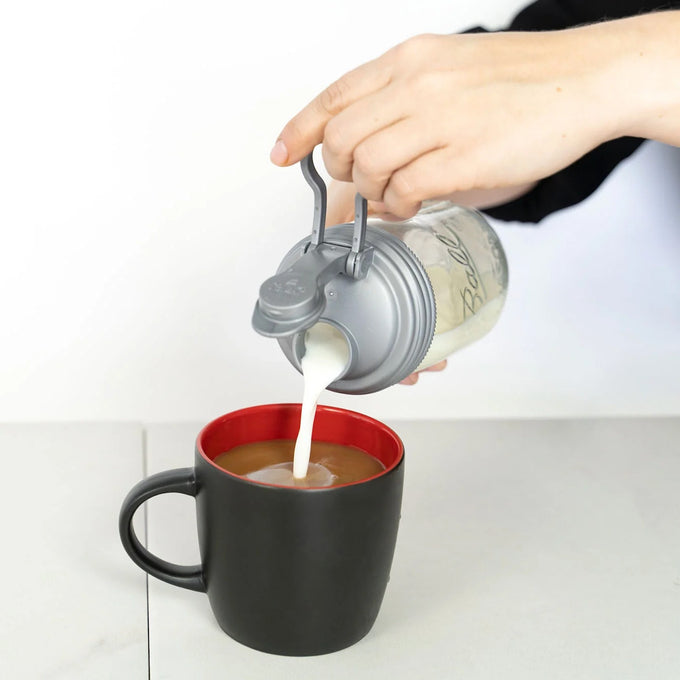 Hand pouring milk from a clear glass jar with a gray no-break plastic pour lid featuring a flip-top spout and carry loop into a black coffee mug, showcasing durable USA-made kitchen accessories ideal for Kitchen & Food, Dinnerware, Kitchen Gadgets, Kitchen Accessories, Best Sellers, and Promo Eligible collections.