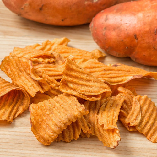 Close-up of rippled sweet potato chips seasoned with sea salt on a wooden surface with whole sweet potatoes in the background, part of gluten free USA made snacks and best sellers collections.