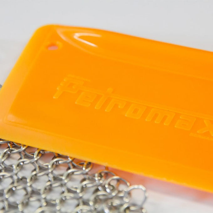 Close-up of bright orange pan scraper with embossed logo and attached metal chain, designed for gentle cleaning of cast iron cookware, featured in Kitchen & Food, Cookware, Cast Iron, Best Sellers, and Promo Eligible collections.