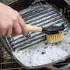 Scrubbing brush with stiff nylon bristles and ergonomic rubberwood handle cleaning a soapy cast iron grill pan, suited for Kitchen & Food, Cookware, Cast Iron, Best Sellers, and Promo Eligible collections.