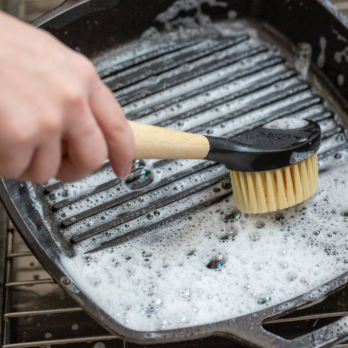 Scrubbing brush with stiff nylon bristles and ergonomic rubberwood handle cleaning a soapy cast iron grill pan, suited for Kitchen & Food, Cookware, Cast Iron, Best Sellers, and Promo Eligible collections.