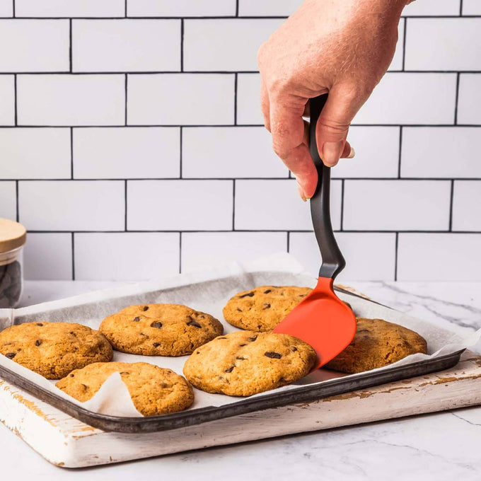 Hand using a flexible spatula with a red blade and black handle to lift freshly baked chocolate chip cookies on a parchment-lined baking sheet, perfect for Kitchen & Food and Kitchen Gadgets collections.