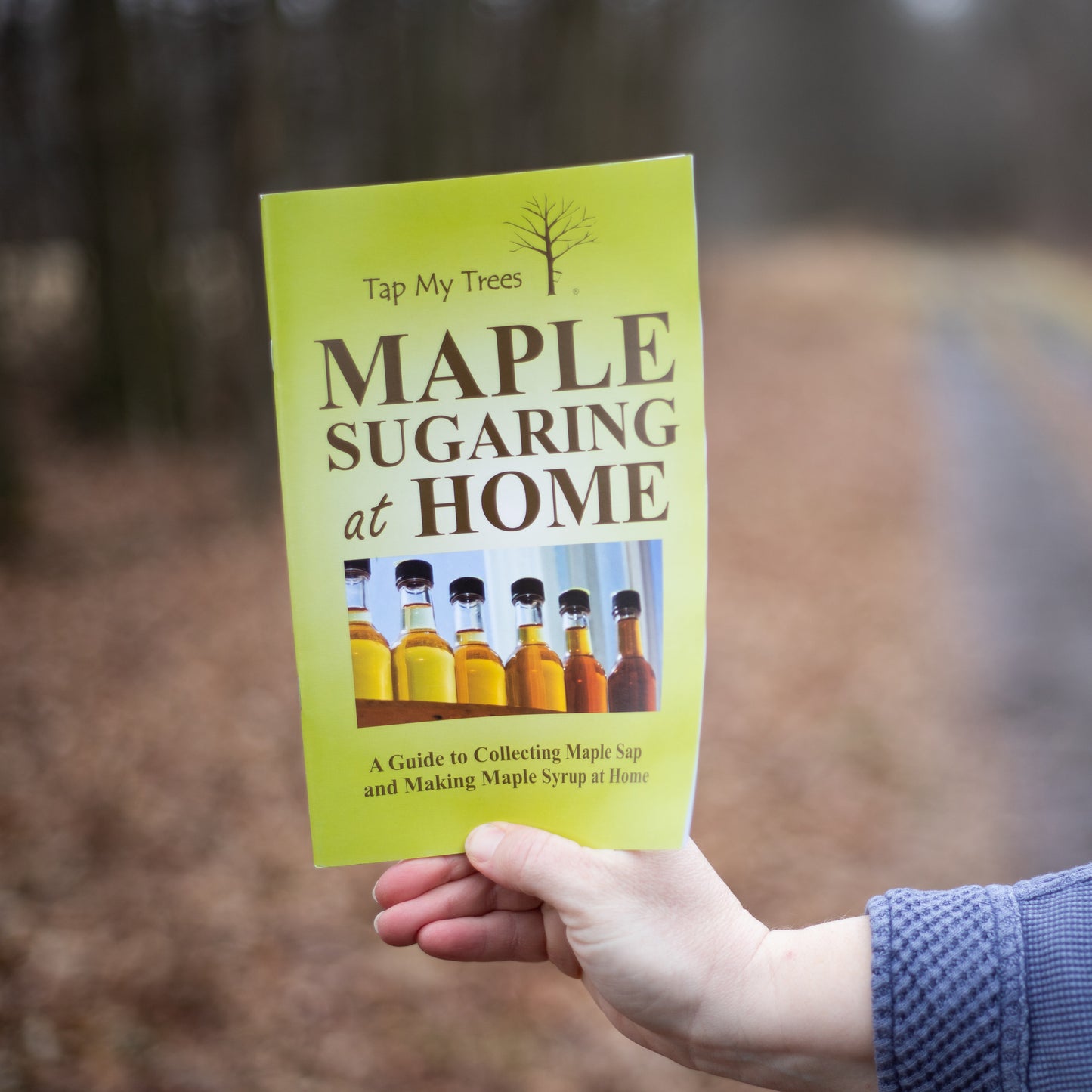 Person holding a yellow booklet titled "Maple Sugaring at Home" outdoors, with a blurred natural background of bare trees and a forest path, part of Farm, Garden & Tool and Orchard collections.