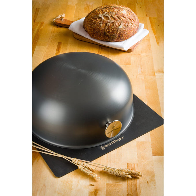 Matte black Brod & Taylor baking shell with smooth dome and metal knob on a black mat, accompanied by crusty artisan boule bread and wheat stalks, suited for Kitchen & Food, Cookware, Baking Supplies, and Bread Making collections.