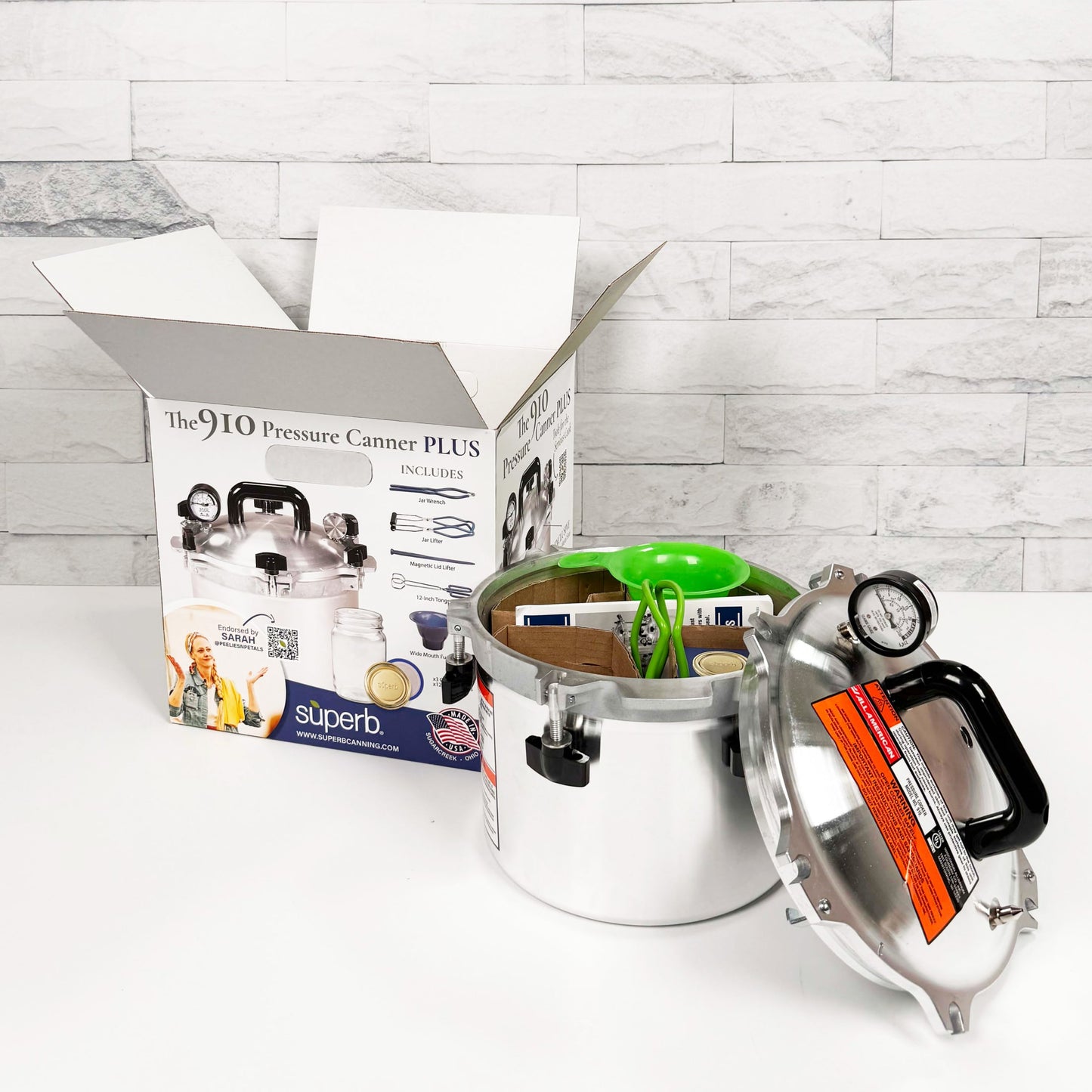 Stainless steel pressure canner with pressure gauge and black handle, shown with lid off and packing box in background, ideal for food preservation and kitchen use, made in USA.