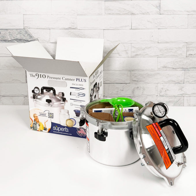 Stainless steel pressure canner with pressure gauge and black handle, shown with lid off and packing box in background, ideal for food preservation and kitchen use, made in USA.