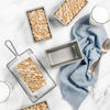 Set of four steel mini loaf pans with Americoat finish, shown with freshly baked bread drizzled with icing, styled with vintage silver forks, light blue linen, and glasses of milk, ideal for kitchen and baking supplies collections emphasizing USA made cookware.