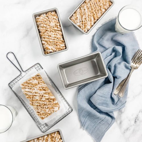Set of four steel mini loaf pans with Americoat finish, shown with freshly baked bread drizzled with icing, styled with vintage silver forks, light blue linen, and glasses of milk, ideal for kitchen and baking supplies collections emphasizing USA made cookware.