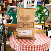 50-pound bag of Hard Red Winter Wheat grain from a Montana farm, placed on a red and white checkered tablecloth with vintage green grain milling equipment in a rustic shop, ideal for Farm, Garden & Tool, Kitchen & Food, Emergency Supplies, Pantry, and Bread Making collections.