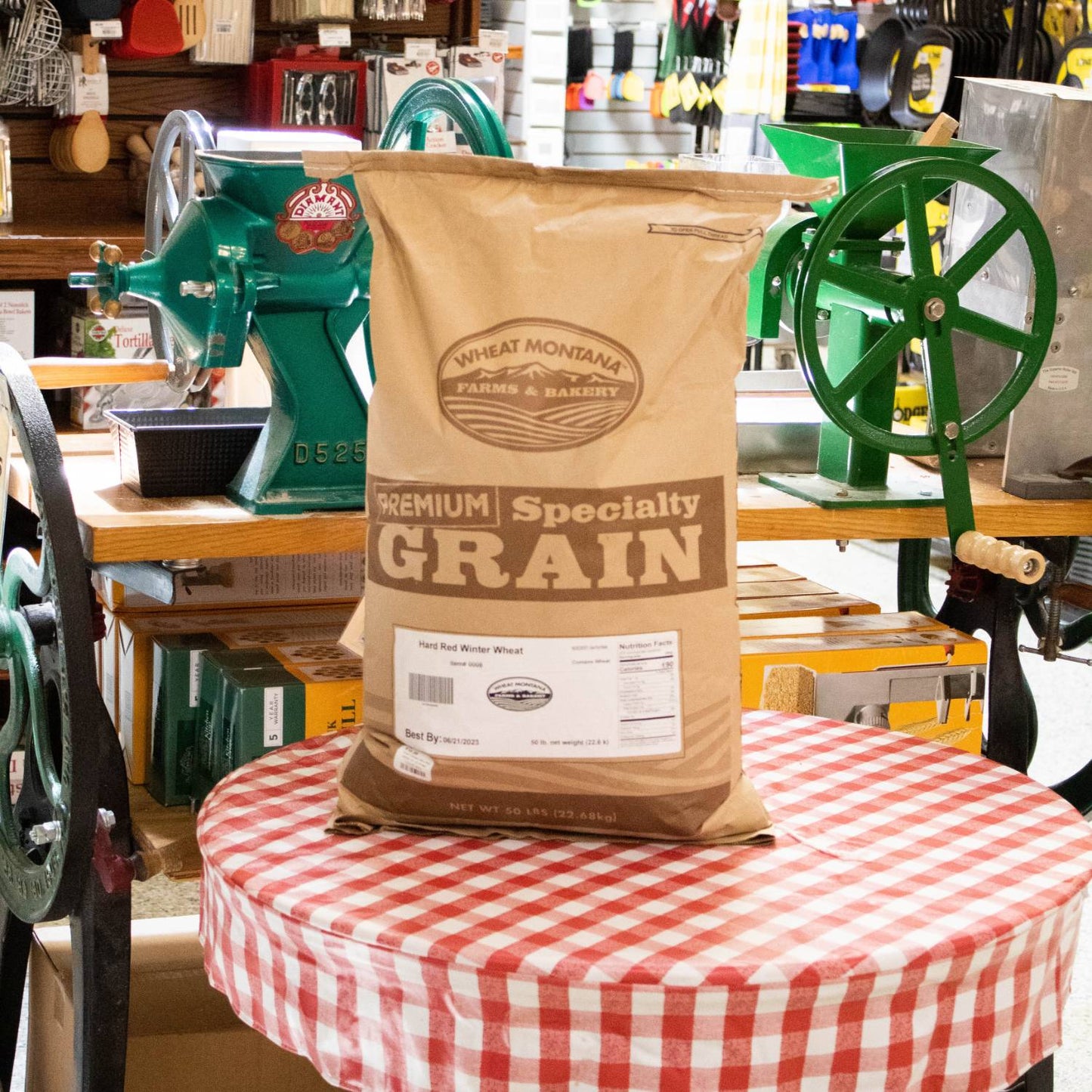 50-pound bag of Hard Red Winter Wheat grain from a Montana farm, placed on a red and white checkered tablecloth with vintage green grain milling equipment in a rustic shop, ideal for Farm, Garden & Tool, Kitchen & Food, Emergency Supplies, Pantry, and Bread Making collections.