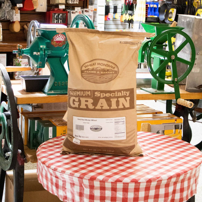 50-pound bag of Hard Red Winter Wheat grain from a Montana farm, placed on a red and white checkered tablecloth with vintage green grain milling equipment in a rustic shop, ideal for Farm, Garden & Tool, Kitchen & Food, Emergency Supplies, Pantry, and Bread Making collections.