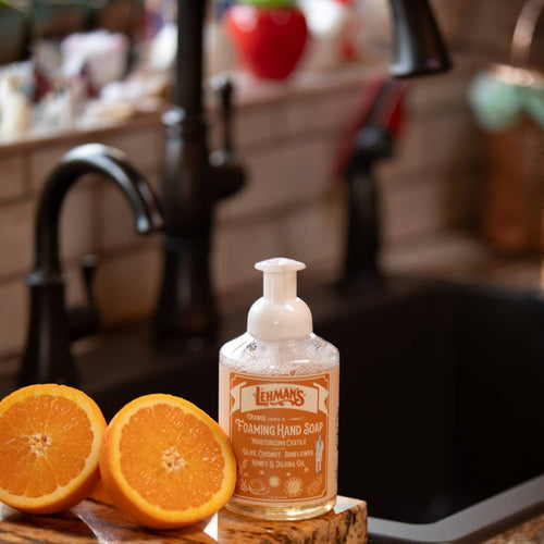 Handcrafted foaming hand soap with natural moisturizing oils including olive, coconut, sunflower, honey, and jojoba, presented next to fresh orange halves near a kitchen sink, fitting Health & Wellness, Grooming and Personal Hygiene, Soaps and Soapmaking, USA Made, and Best Sellers collections.