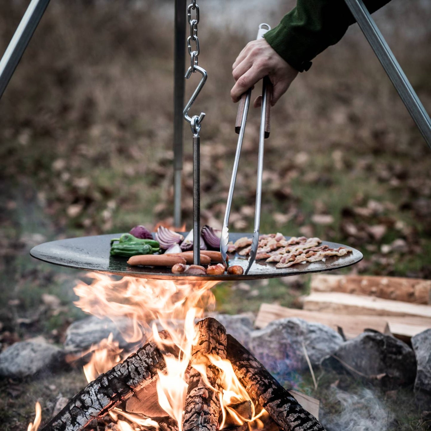 Brushed stainless steel tongs with a practical locking mechanism, used for safely handling food cooking on an outdoor hanging grill over an open fire, ideal for camping and grilling collections.