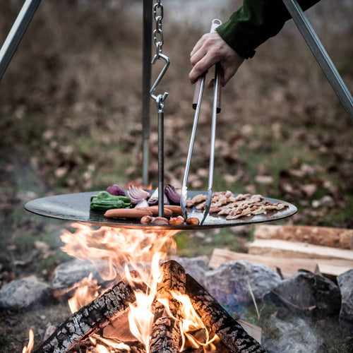 Brushed stainless steel tongs with a practical locking mechanism, used for safely handling food cooking on an outdoor hanging grill over an open fire, ideal for camping and grilling collections.