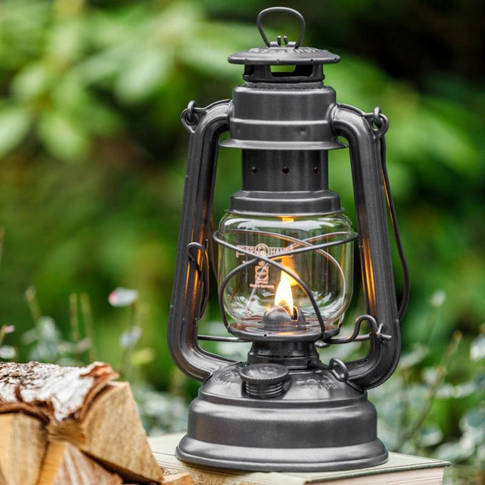 Handcrafted dark galvanized oil lantern with thermal heat-resistant globe glowing warmly outdoors on a natural wood surface, perfect for Lamps & Lighting, Camping, and Emergency Supplies collections.