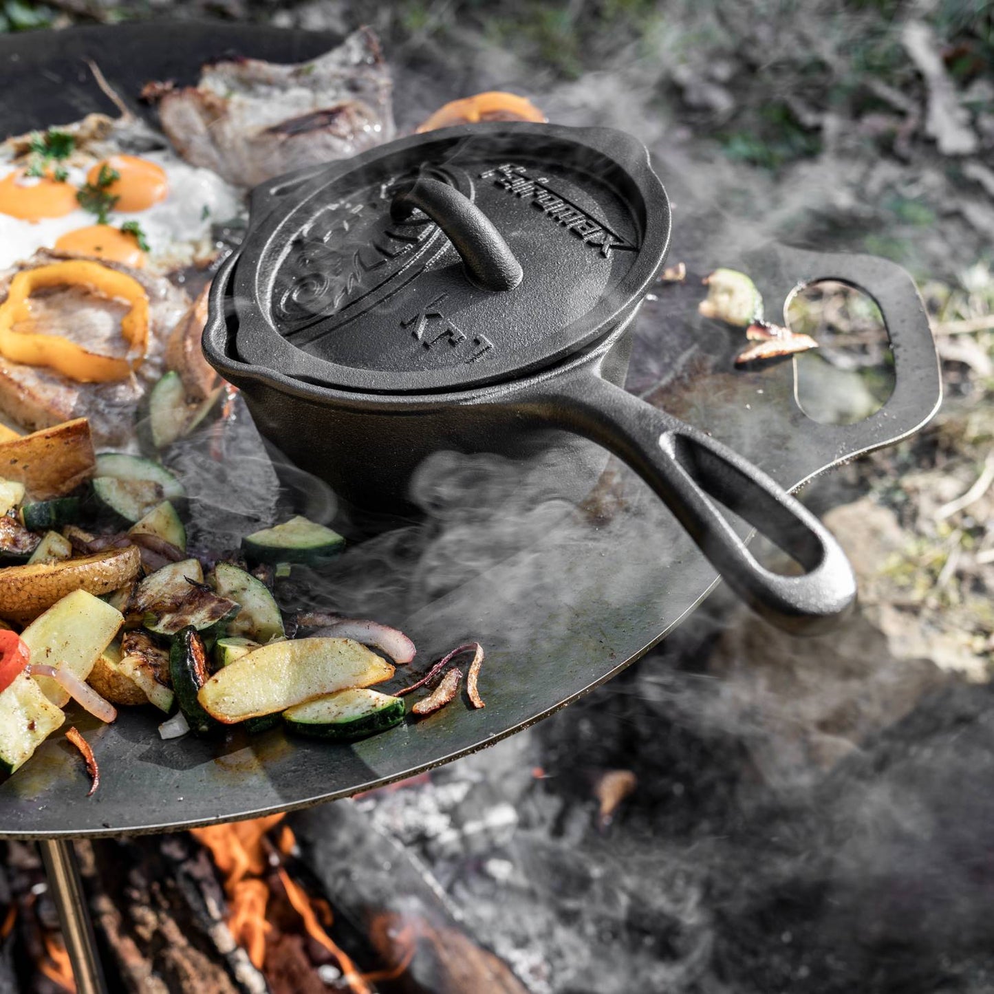 Handcrafted cast iron saucepan with lid and side pouring spouts, cooking over open fire with vegetables and eggs, ideal for farm, garden, camping, and outdoor cookware collections.