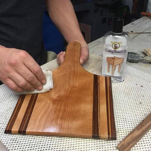 Hand applying clear mineral oil to a striped wooden cutting board with a cloth, shown alongside a bottle of cutting board oil, highlighting wood care in Kitchen & Food and USA Made collections.