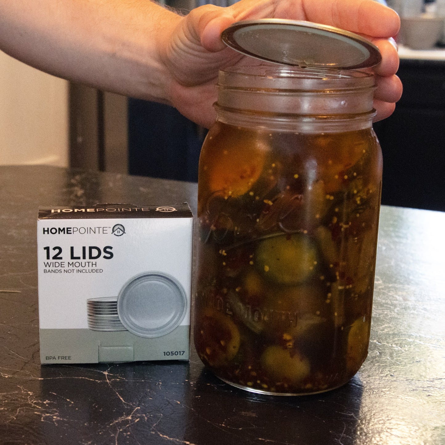 Hand holding a silver metal wide-mouth canning lid above a glass jar filled with pickled cucumbers, next to a box of 12 BPA-free canning lids for Kitchen & Food, Food Preservation, and Canning collections.