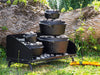 Heavy-duty black steel outdoor cooking table with multiple cast iron Dutch ovens stacked and heated with charcoal briquettes, set on grass near tree trunk, ideal for camping and garden use.
