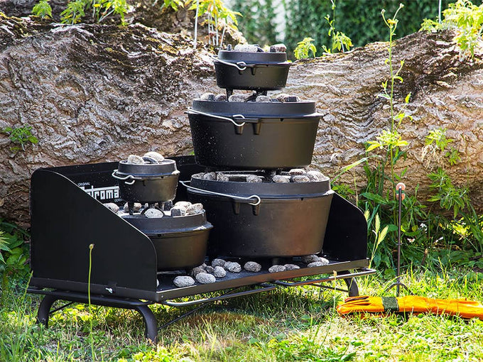 Heavy-duty black steel outdoor cooking table with multiple cast iron Dutch ovens stacked and heated with charcoal briquettes, set on grass near tree trunk, ideal for camping and garden use.
