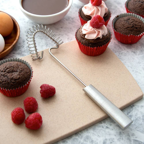 Stainless steel coil whisk with a long handle resting on a cutting board beside chocolate cupcakes topped with raspberry frosting and fresh raspberries, ideal for Kitchen & Food and Cooking and Baking Helpers collections.