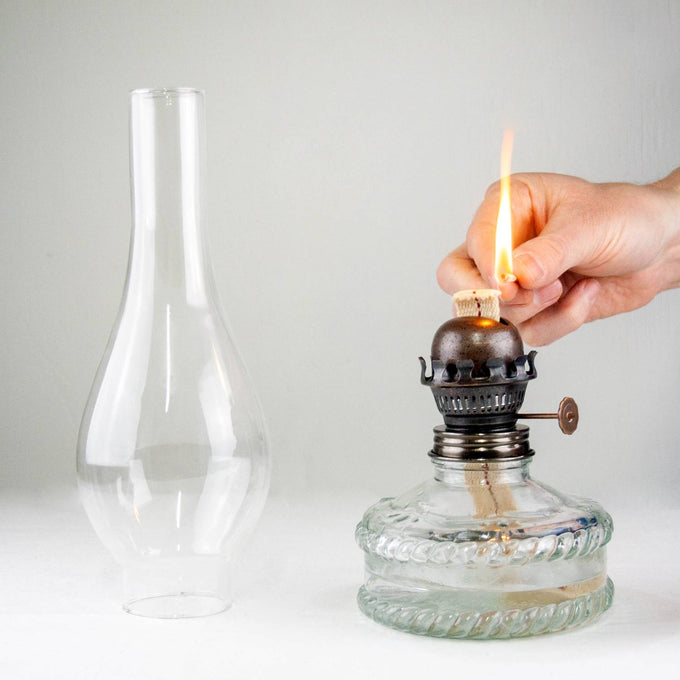 Hand turning wick knob on a handcrafted oil lamp with a clear glass base and fabric wick, shown alongside its tall glass chimney, ideal for Lamps & Lighting and Oil Lamps collections.