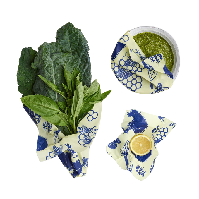 Set of three reusable beeswax food wraps in cream with blue bee and honeycomb designs, covering fresh greens, a bowl of green sauce, and a halved lemon, eco-friendly and handmade in USA collections.