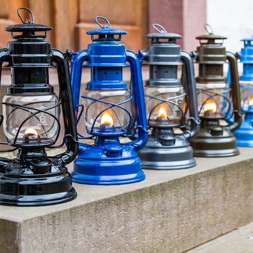 Handcrafted galvanized oil lanterns in black, blue, gray, and olive tones, some softly glowing, displayed in a row outdoors, ideal for Lamps & Lighting, Camping, and Emergency Supplies collections.