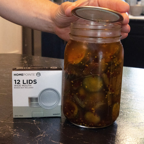 Hand holding wide-mouth lid above a glass jar filled with homemade pickles, next to a box of 12 BPA-free sealing lids, suited for Kitchen & Food, Food Preservation, and Canning collections.