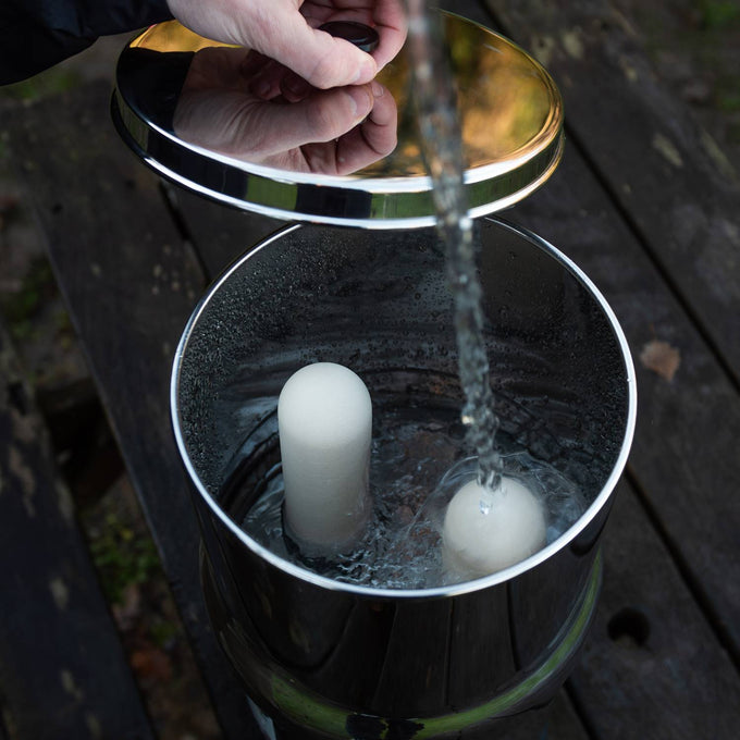 Hand filling a polished stainless steel gravity water filter with visible ceramic filter elements inside, suitable for camping, emergency supplies, and farm collections.
