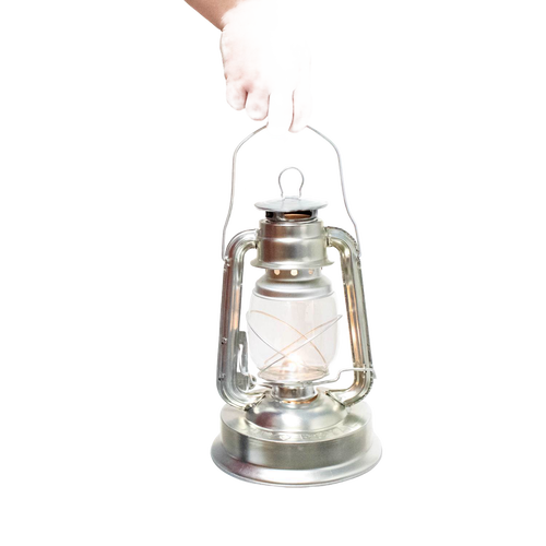 Hand holding a silver metal oil lantern with a clear glass globe and visible wick, designed for outdoor use in camping, pest control, and farm or garden lighting collections.
