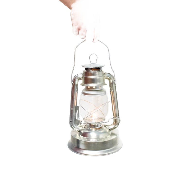 Hand holding a silver metal oil lantern with a clear glass globe and visible wick, designed for outdoor use in camping, pest control, and farm or garden lighting collections.