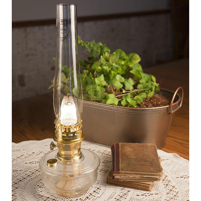 Handcrafted Aladdin Genie III Oil Lamp with clear dimpled glass bowl and polished brass burner, displayed on lace tablecloth beside vintage books and a metal planter, ideal for Farm, Garden & Tool, Lamps & Lighting, and Emergency Supplies collections.
