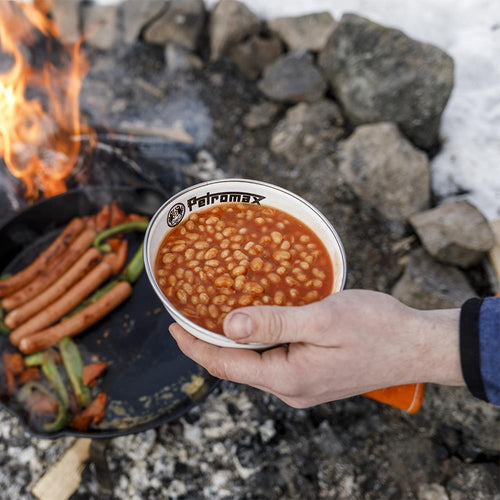 Hand holding a white enamel bowl with black rim and logo filled with baked beans near an outdoor campfire with sausages and vegetables cooking in a cast iron skillet, ideal for Farm, Garden & Tool, Kitchen & Food, Camping, Dinnerware, Best Sellers, and Promo Eligible collections.