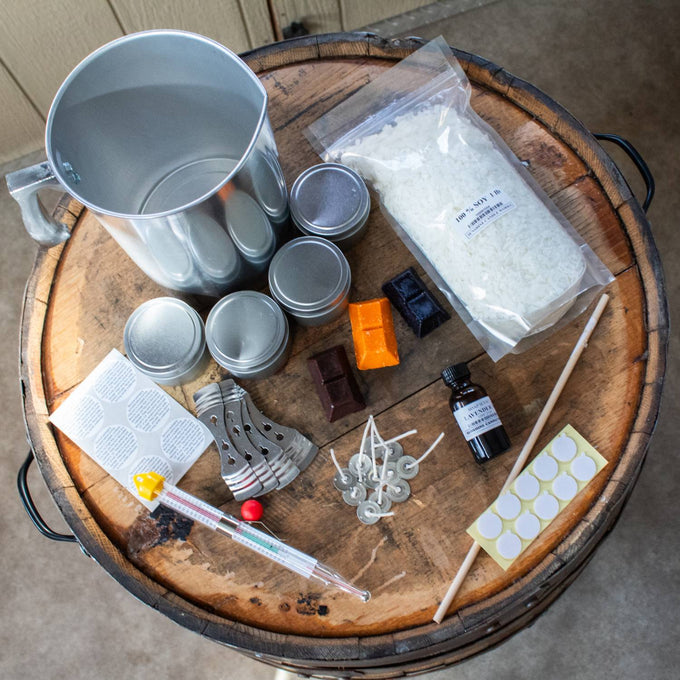 Collection of handcrafted soy candle making supplies including a metal melting pot, soy wax flakes, scented oil, wick sustainers, dye chips, wick stickers, and wicks on a rustic wooden surface, ideal for beginners in lamps and candlemaking supplies.